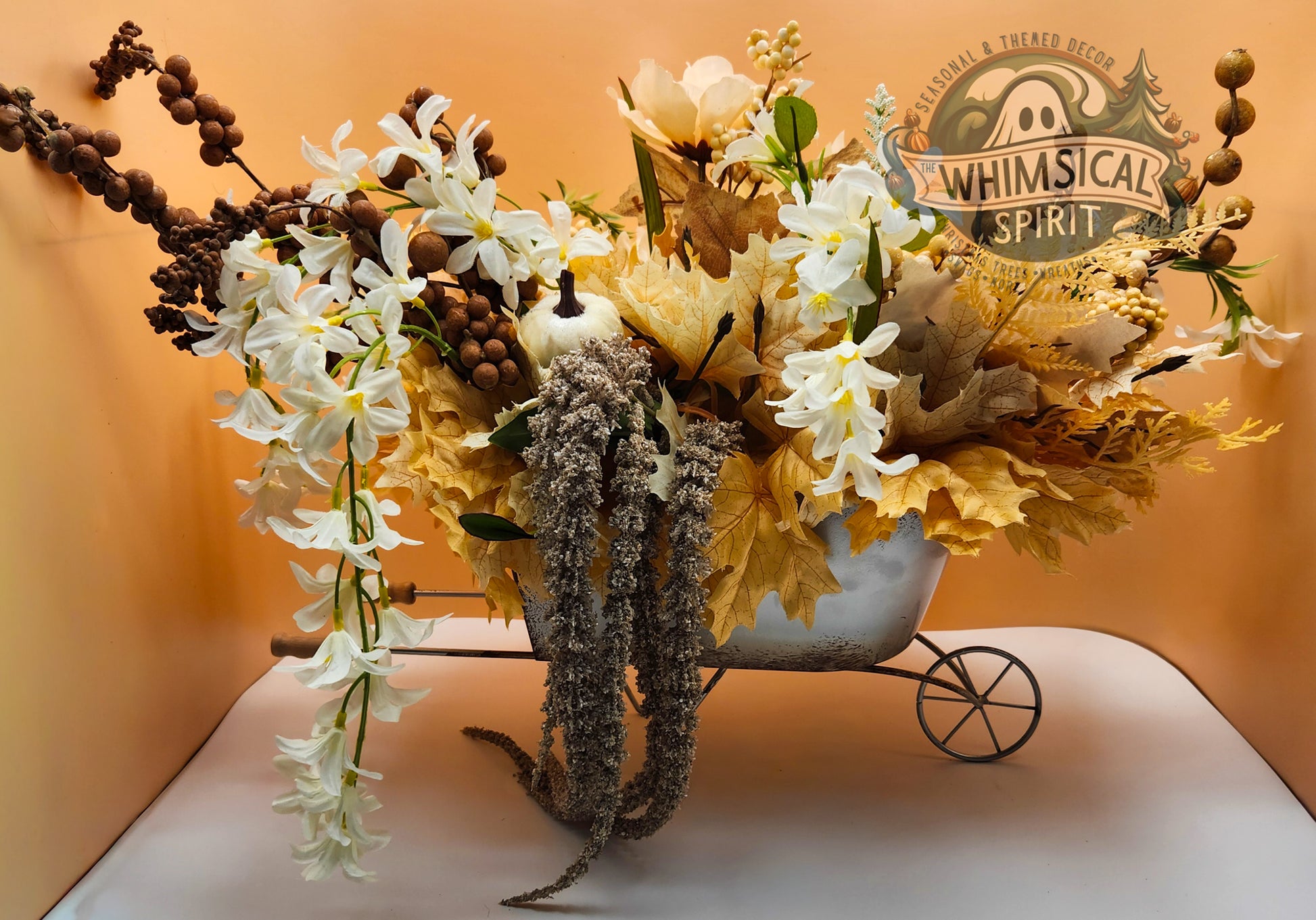 Decorative flower arrangement in a small wheelbarrow with 'Whimsical Spirit' branding on an orange background.