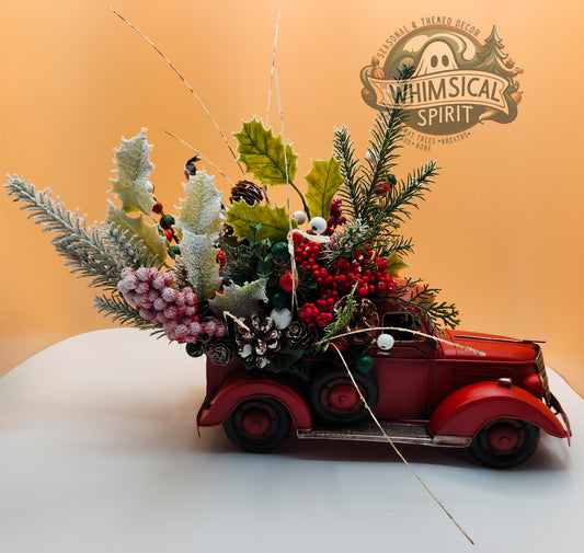 Christmas floral arrangement with greenery in a red vintage car model, featuring The Whimsical Spirit logo in a festive holiday display.