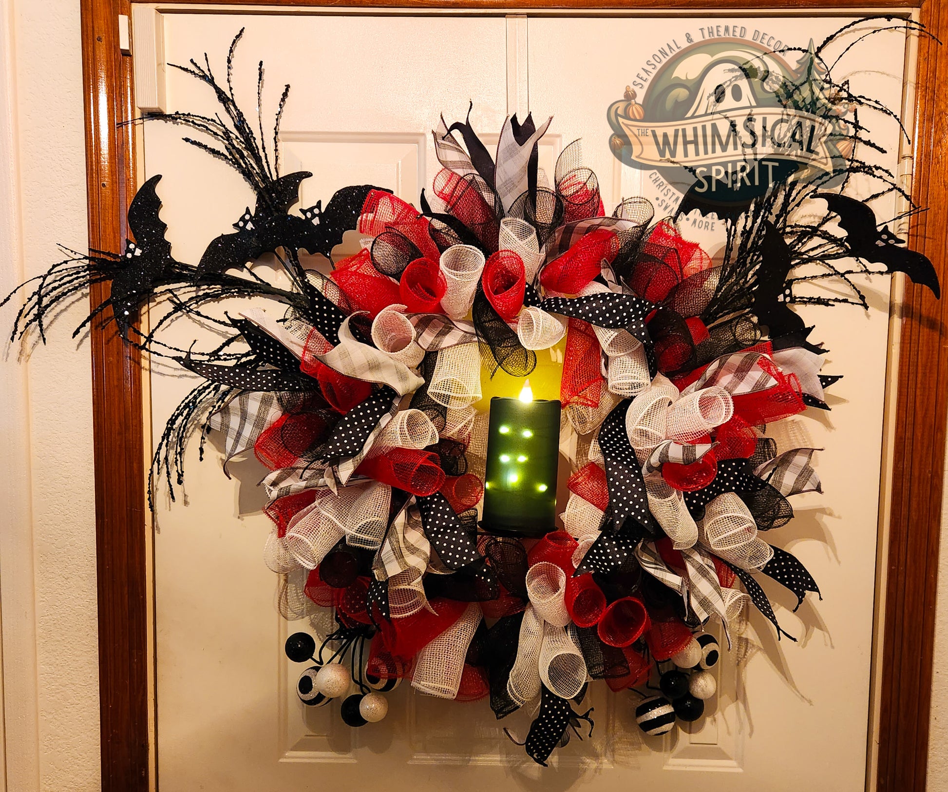 Halloween Decorative wreath with red, white, and black ribbons  and black glittered bats on a door with 'Whimsical Spirit' sign.