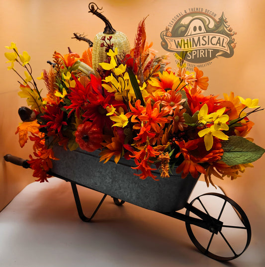 Decorative wheelbarrow filled with autumn-themed flowers and pumpkins, featuring 'Whimsical Spirit' logo.