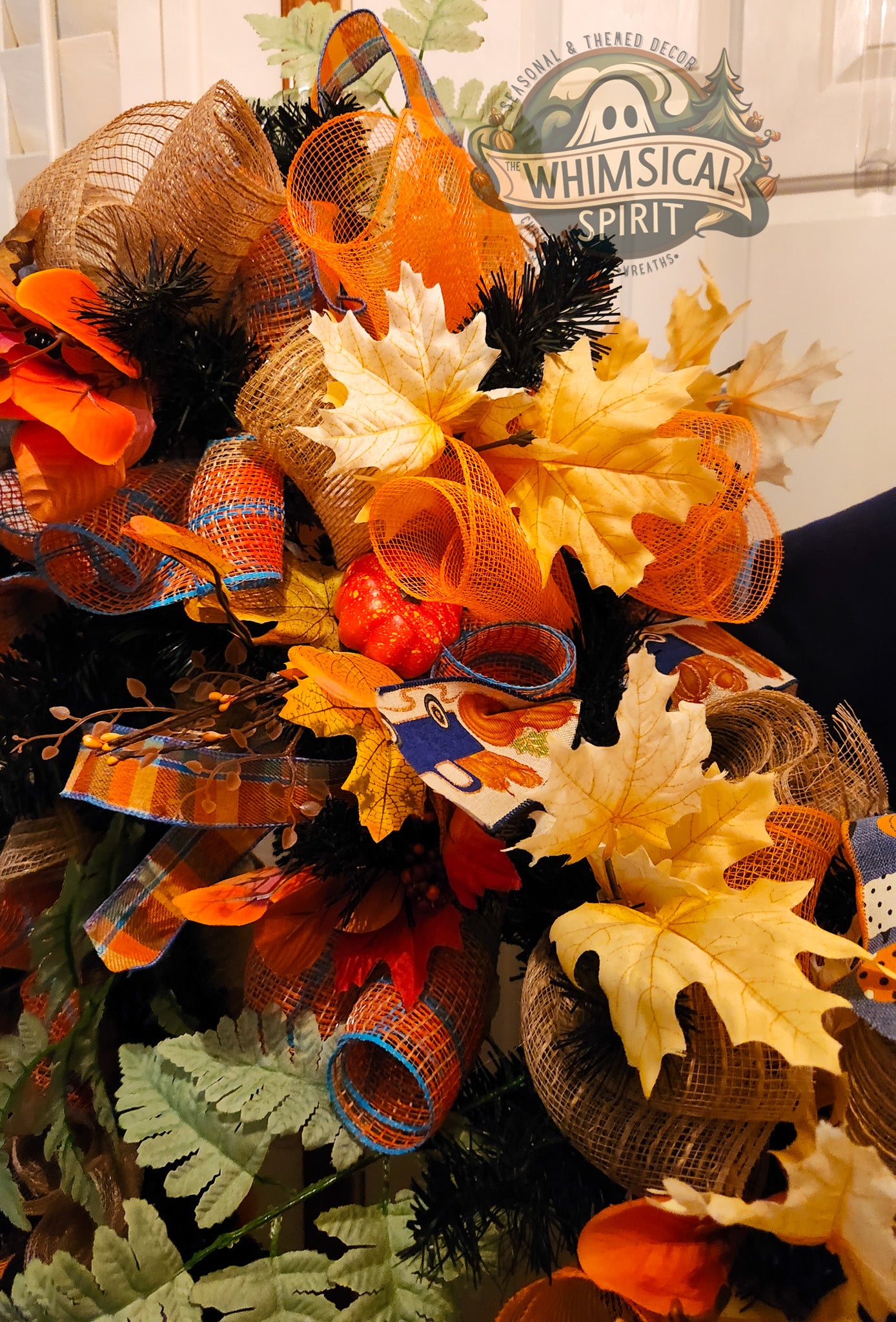 Decorative wreath with a fall therme with orange, yellow, and black ribbons and leaves, featuring a 'Whimsical Spirit' sign in the background.
