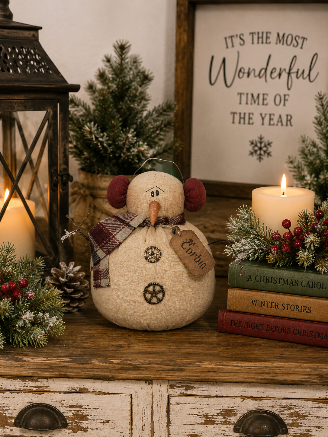 Primitive snowman soft sculpture styled on a rustic tabletop with Christmas greenery, candles, pinecones, lantern, and festive holiday decor.