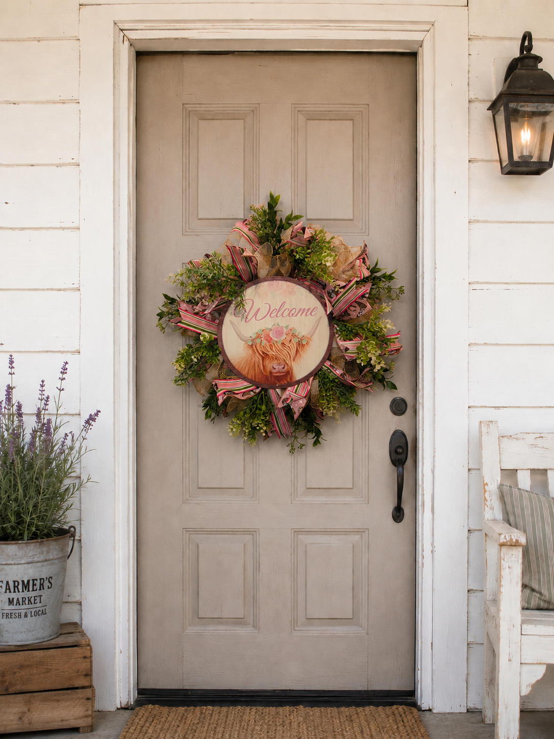 Handmade highland cow welcome wreath styled on a farmhouse front door with greenery, pink ribbon, burlap accents, and rustic porch decor.