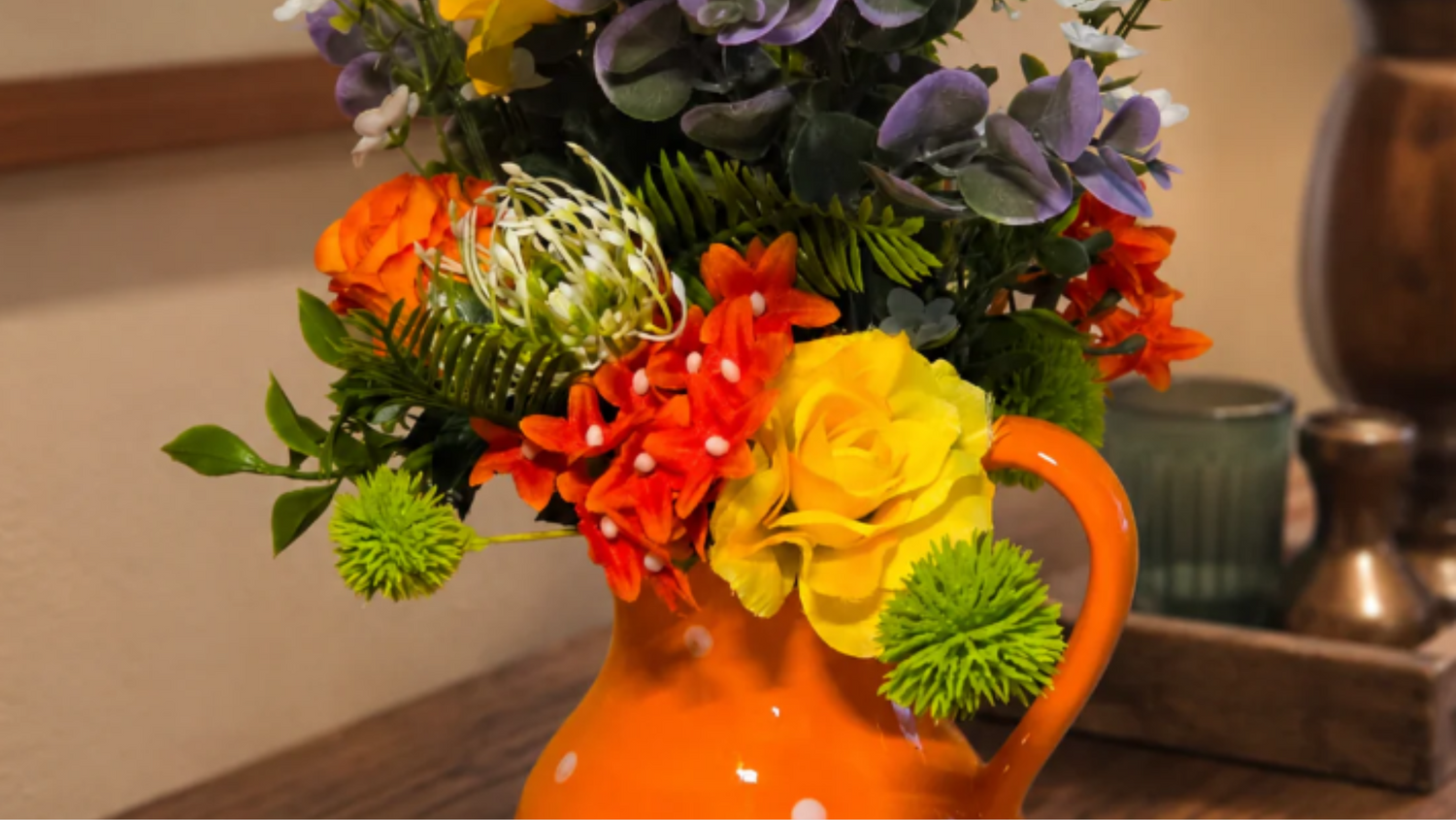 Handmade floral centerpiece in an orange pitcher with yellow, orange, purple, and green flowers for cheerful seasonal table decor.