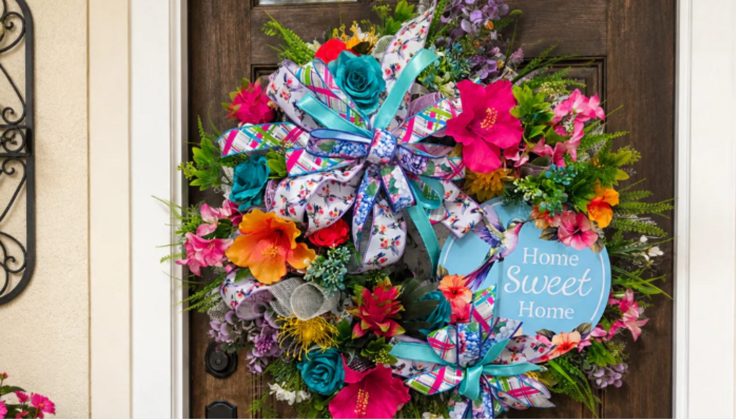 Handmade floral wreath with bright pink, teal, orange, and purple flowers, layered ribbon, greenery, and a Home Sweet Home sign on a front door.