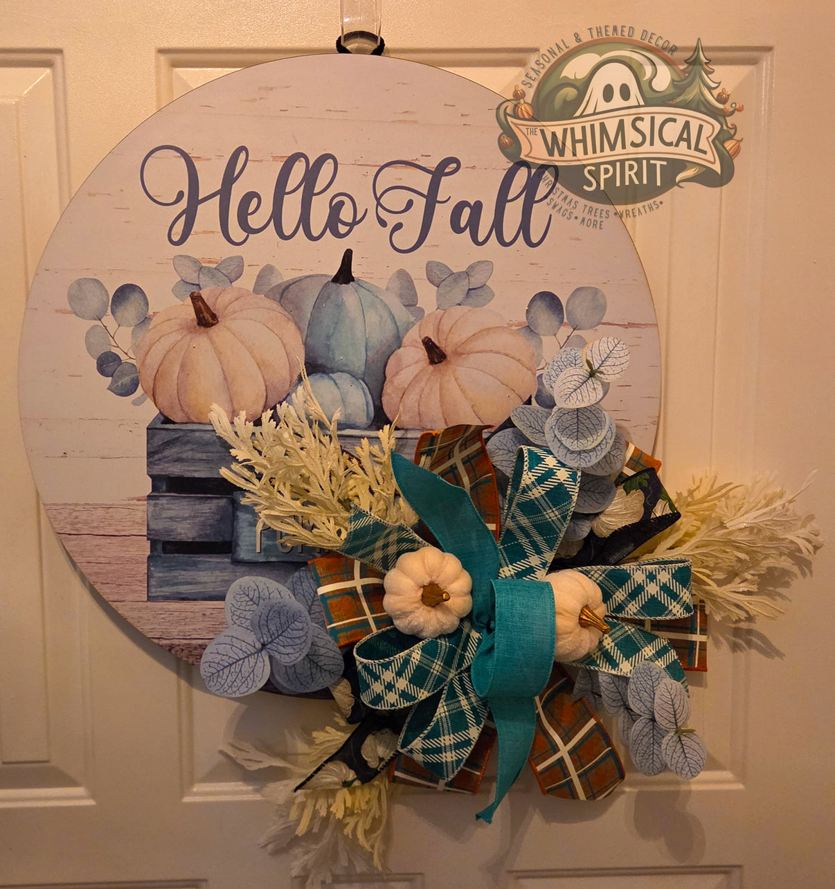 Round Hello Fall pumpkin door hanger displayed on a white paneled door, featuring soft blue and cream pumpkins, eucalyptus-style leaves, and a layered teal, plaid, and rust-toned bow with small cream pumpkins and ivory foliage.