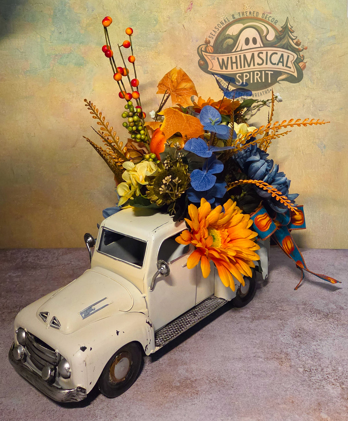 Handmade white vintage truck fall centerpiece filled with blue and orange flowers, pumpkins, berries, leaves, and ribbon.