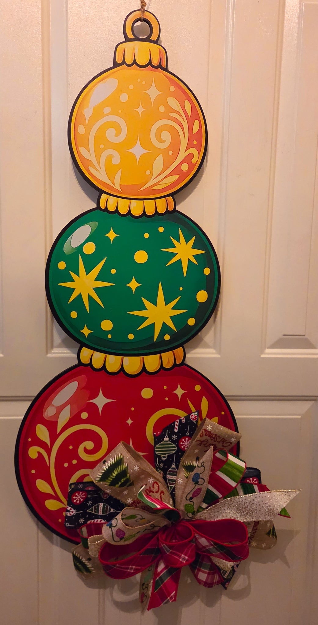 Close-up of the Baubles Full of Cheer door hanger featuring three stacked Christmas ornaments in gold, green, and red with whimsical swirls and starburst details.