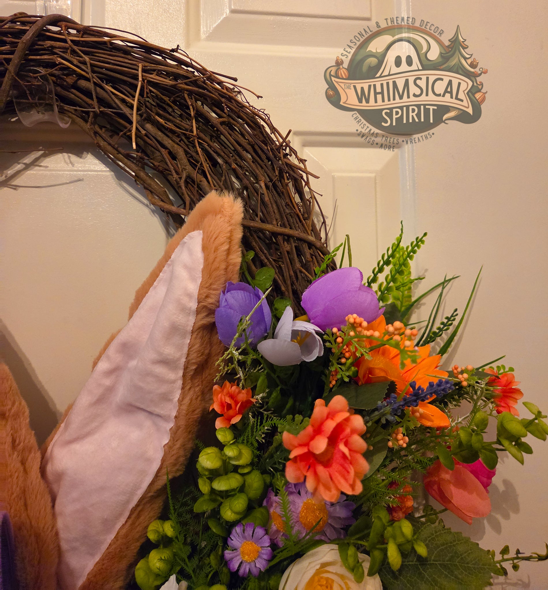 Decorative wreath with flowers and a teddy bear headband on a door, featuring 'Whimsical Spirit' branding.