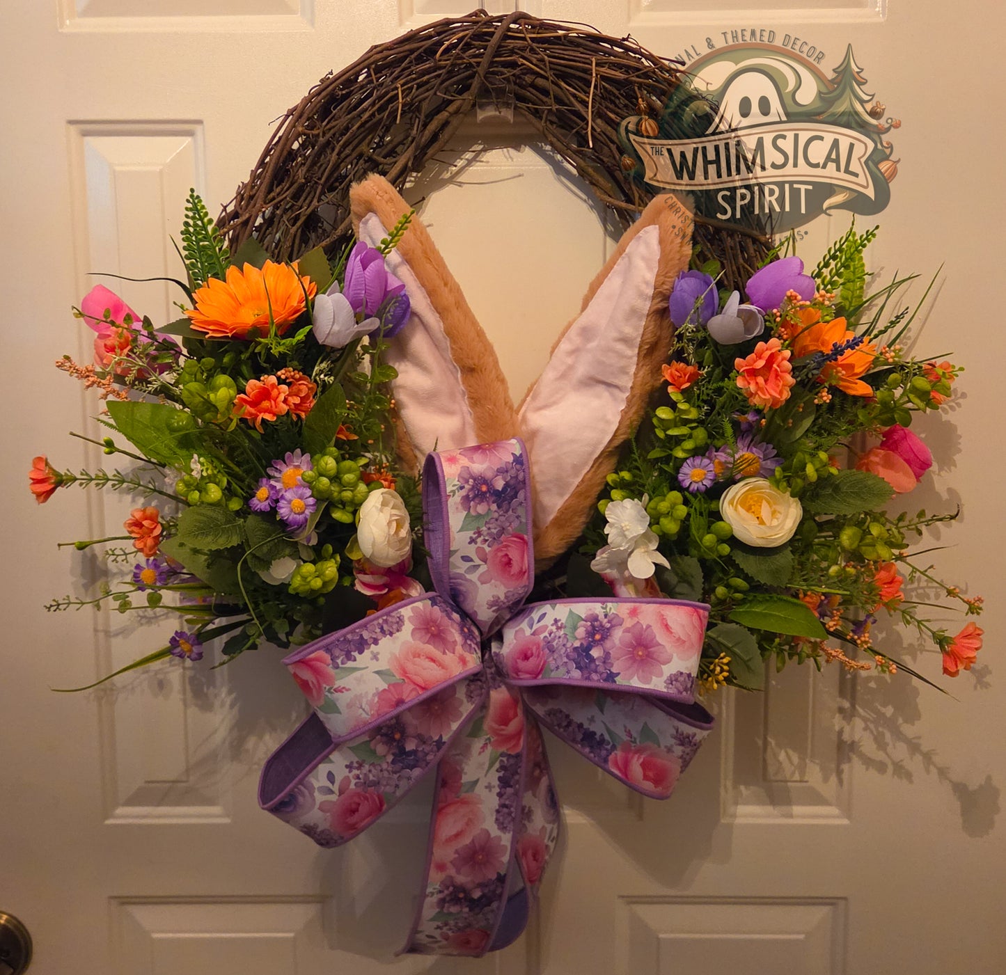Decorative wreath with flowers and bunny ears on a door, branded 'Whimsical Spirit'.