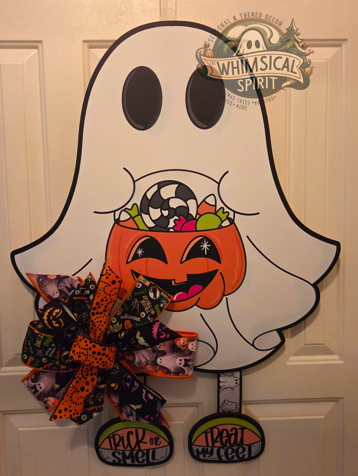 Ghost trick-or-treat Halloween door hanger featuring a smiling pumpkin candy bucket, colorful sweets, a layered orange and black Halloween bow, and dangling “Trick or Treat, Smell My Feet” shoes.