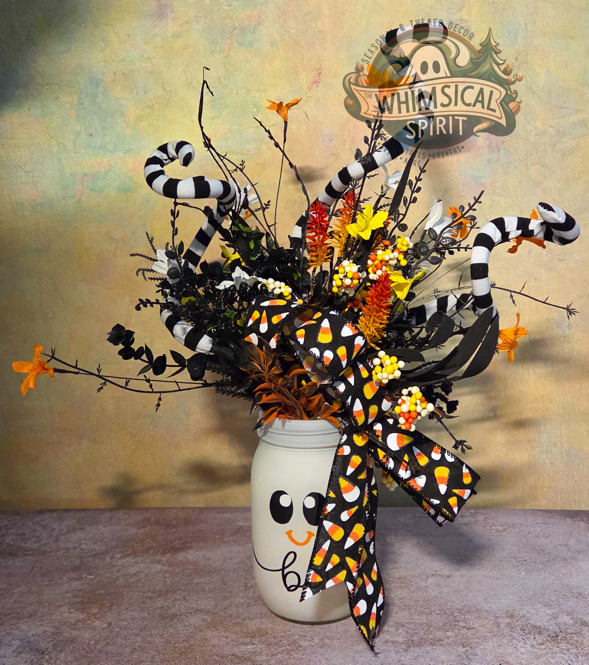 Handmade ghost mason jar centerpiece with candy corn ribbon, striped curls, black foliage, berries, and orange and yellow florals.