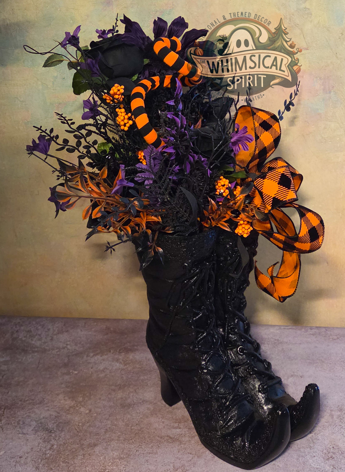 Handmade Halloween centerpiece featuring glossy black witch boots, purple and black florals, striped accents, orange berries, and a large orange plaid bow.