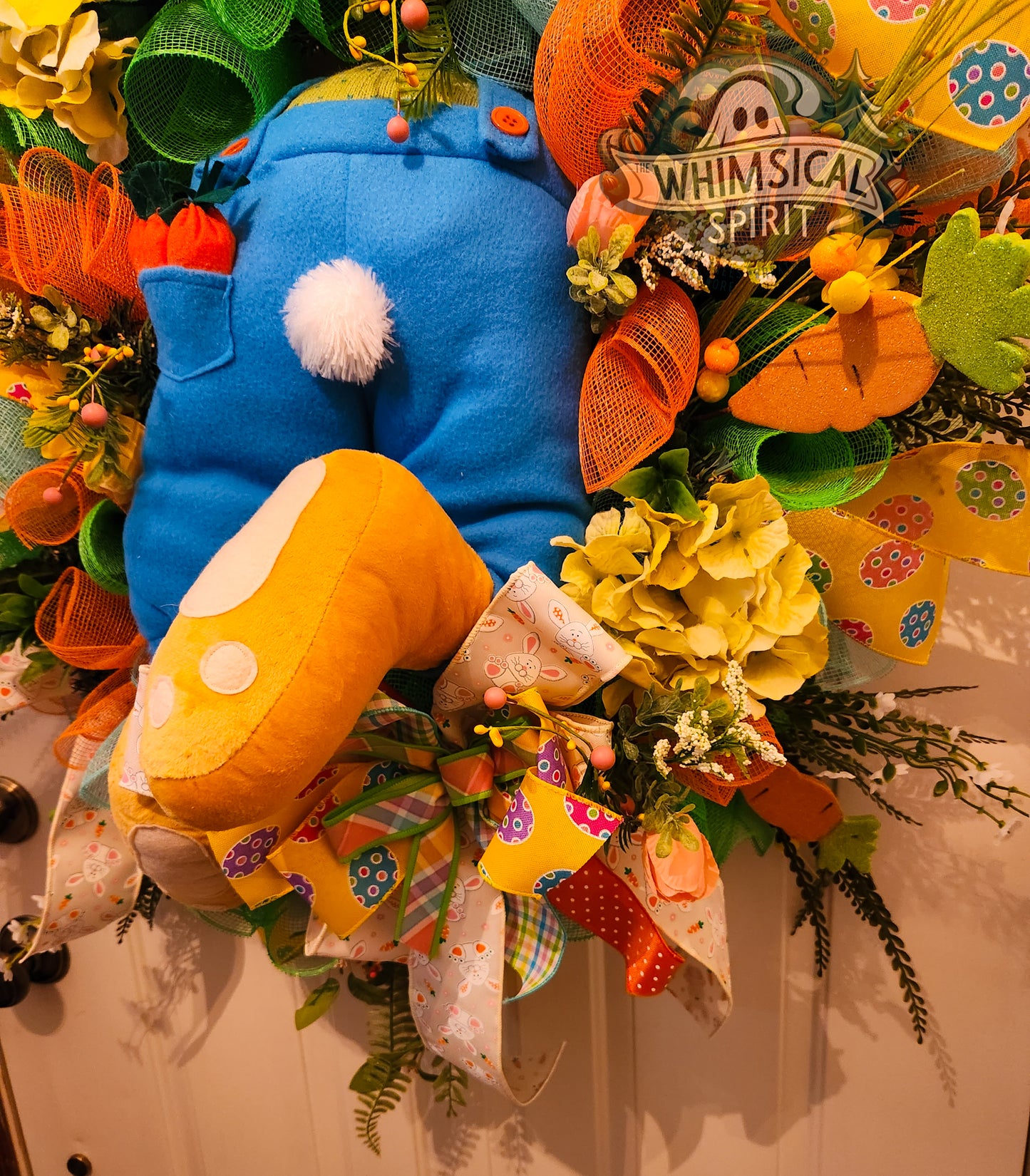 Decorative wreath with a blue and yellow cat plush toy, flowers, and 'Whimsical Spirit' sign.