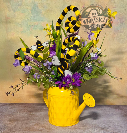 Decorative arrangement with flowers, bees, and a yellow watering can on a textured surface.