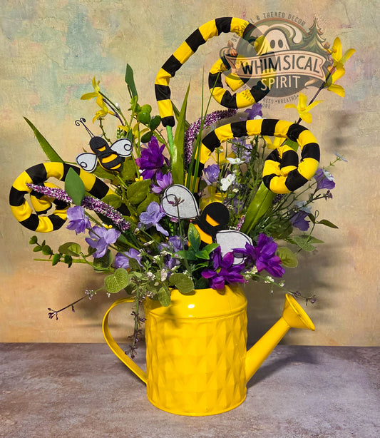 Yellow watering can with floral arrangement and decorative bees against a rustic background