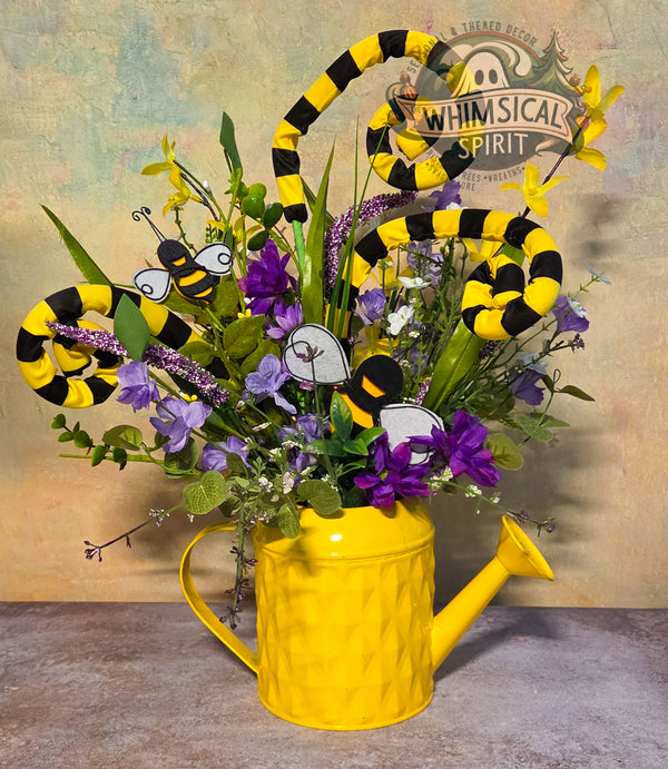 Yellow watering can with floral arrangement and decorative bees against a rustic background