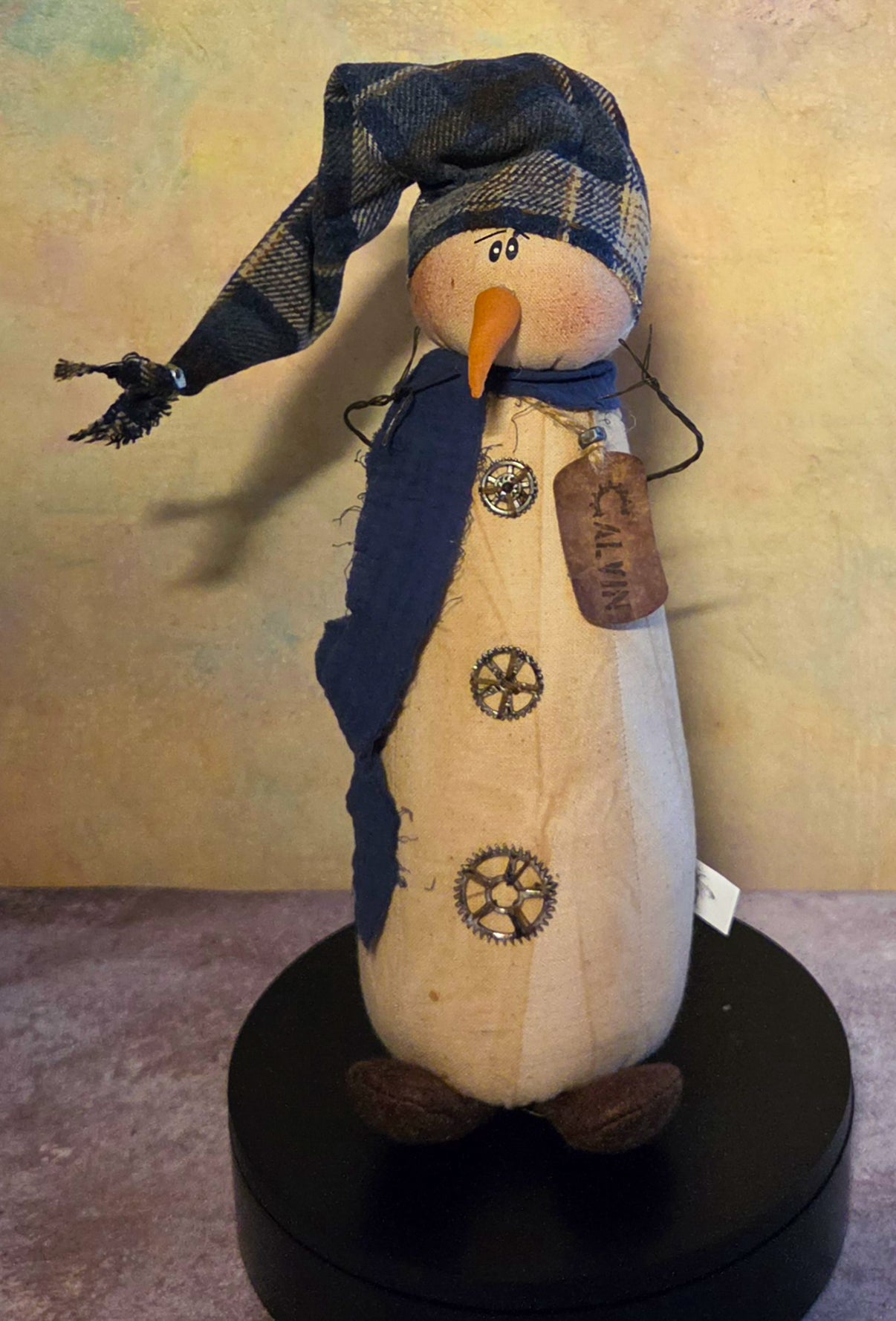 Front view of Calvin snowman soft sculpture with a navy plaid hat, dark blue scarf, carrot nose, twig arms, and gear-style buttons.