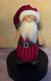 Front view of Chris Santa Claus soft sculpture with a red plaid hat, fluffy white beard, black belt, silver buckle, and dark feet.
