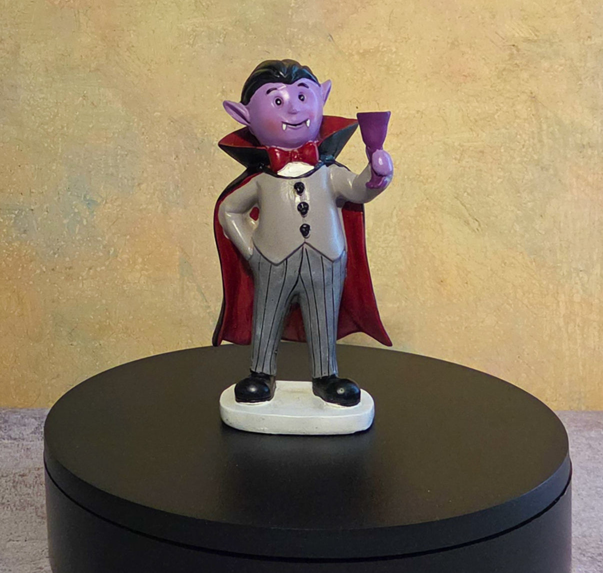 Small purple vampire resin figurine wearing a red-lined cape, cream vest, striped trousers, and black shoes while holding a purple goblet.