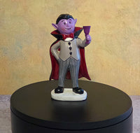 Small purple vampire resin figurine wearing a red-lined cape, cream vest, striped trousers, and black shoes while holding a purple goblet.