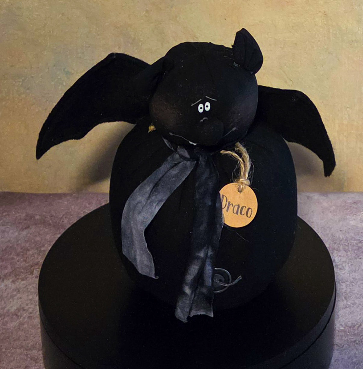 Front view of Draco bat soft sculpture with outstretched wings, pointed ears, charcoal scarf, small fangs, and wooden name tag.