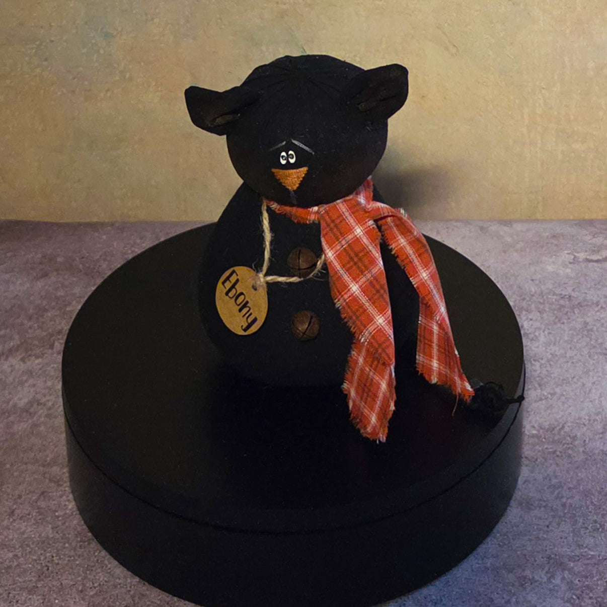 Front view of Ebony black cat soft sculpture with pointed ears, orange plaid scarf, rustic bell buttons, and wooden name tag.