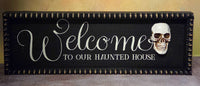 Black framed wooden Halloween sign with white “Welcome to Our Haunted House” lettering and a raised skull accent.