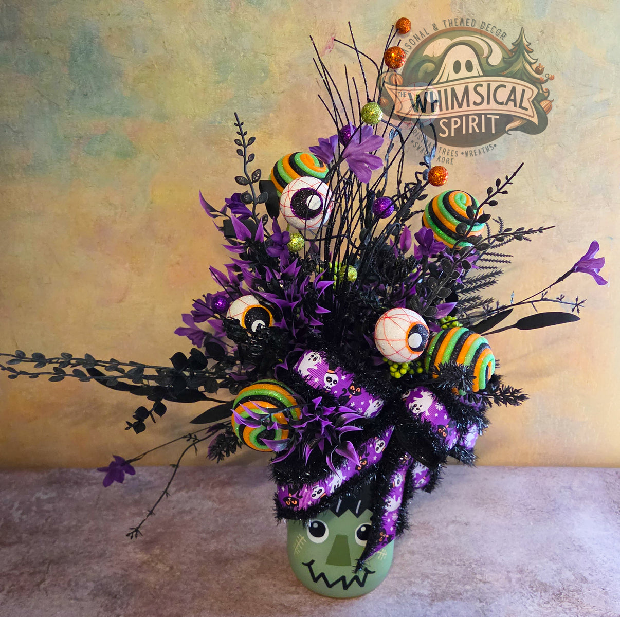 Front view of a Frankenstein Halloween centerpiece with purple flowers, eyeballs, striped candy accents, and a spooky bow.