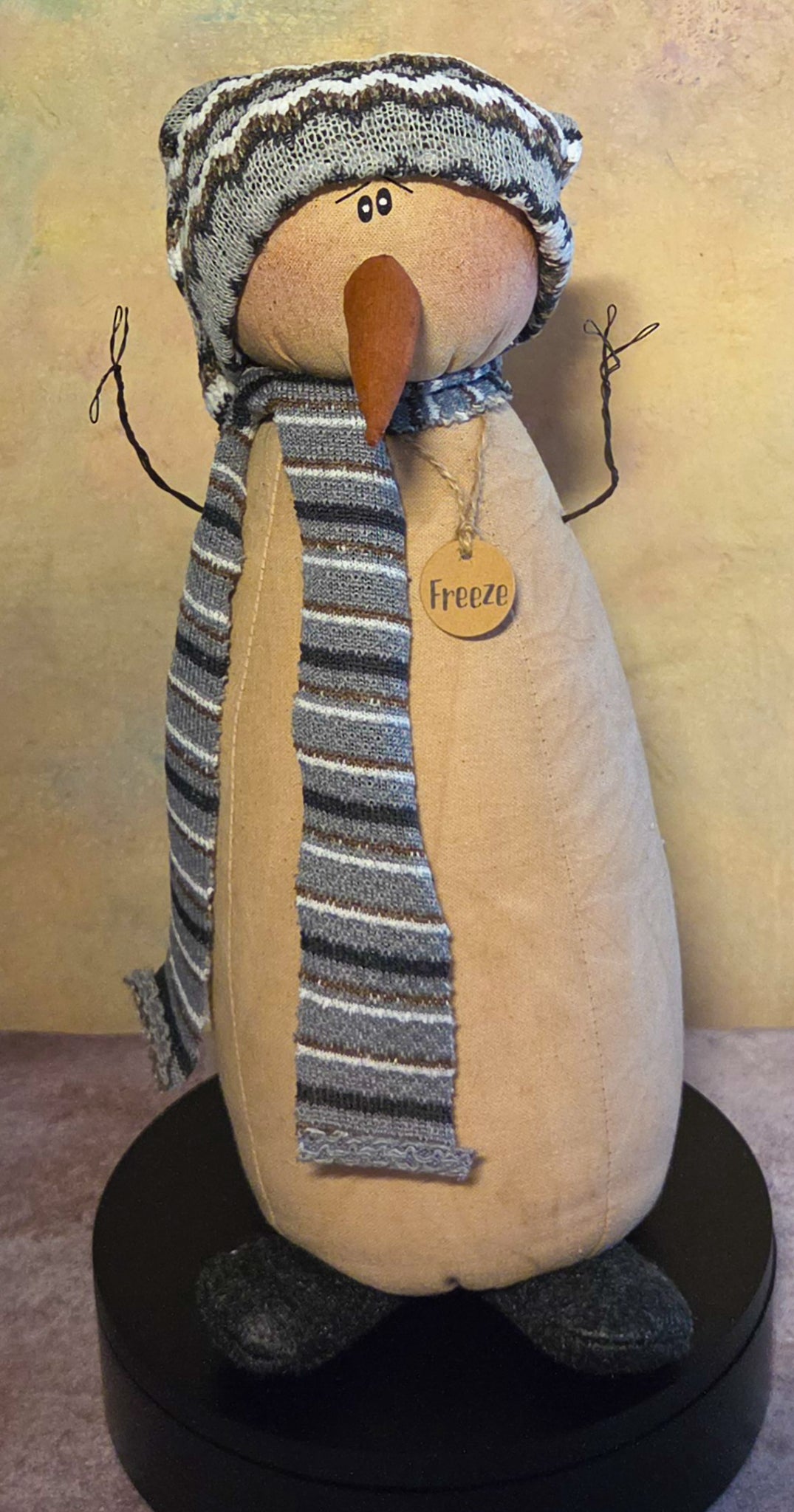 Front view of Freeze snowman soft sculpture with a striped knit hat and scarf, long carrot nose, twig arms, dark feet, and wooden name tag.