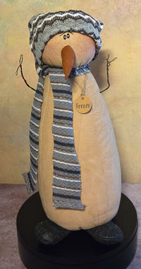 Front view of Freeze snowman soft sculpture with a striped knit hat and scarf, long carrot nose, twig arms, dark feet, and wooden name tag.