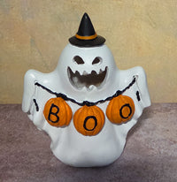 A white ceramic Halloween ghost figurine wearing a black witch hat with an orange band, holding an orange 'BOO' pumpkin banner.
