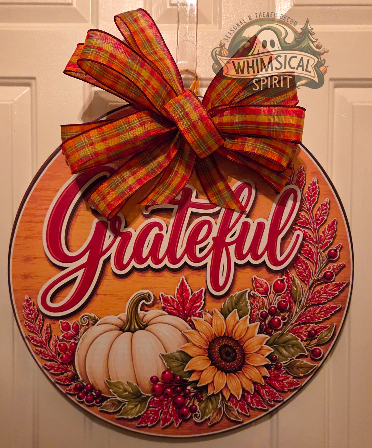Round Grateful fall door hanger with a white pumpkin, sunflower, red leaves and berries, woodgrain background, and large plaid bow.