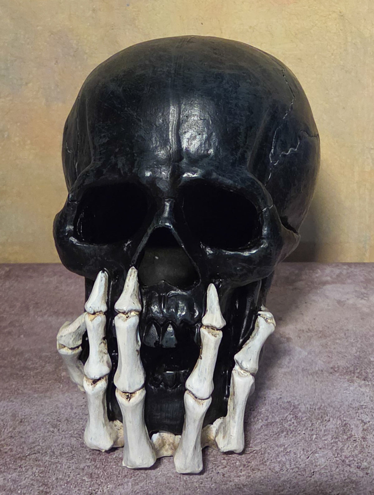 Unlit black skull figurine with dark facial openings and ivory skeletal hands wrapped around its lower jaw.