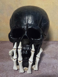 Unlit black skull figurine with dark facial openings and ivory skeletal hands wrapped around its lower jaw.