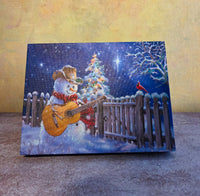 Guitar Snowman light-up Christmas tabletop canvas featuring a cowboy snowman playing guitar beside a glowing Christmas tree and snowy fence.