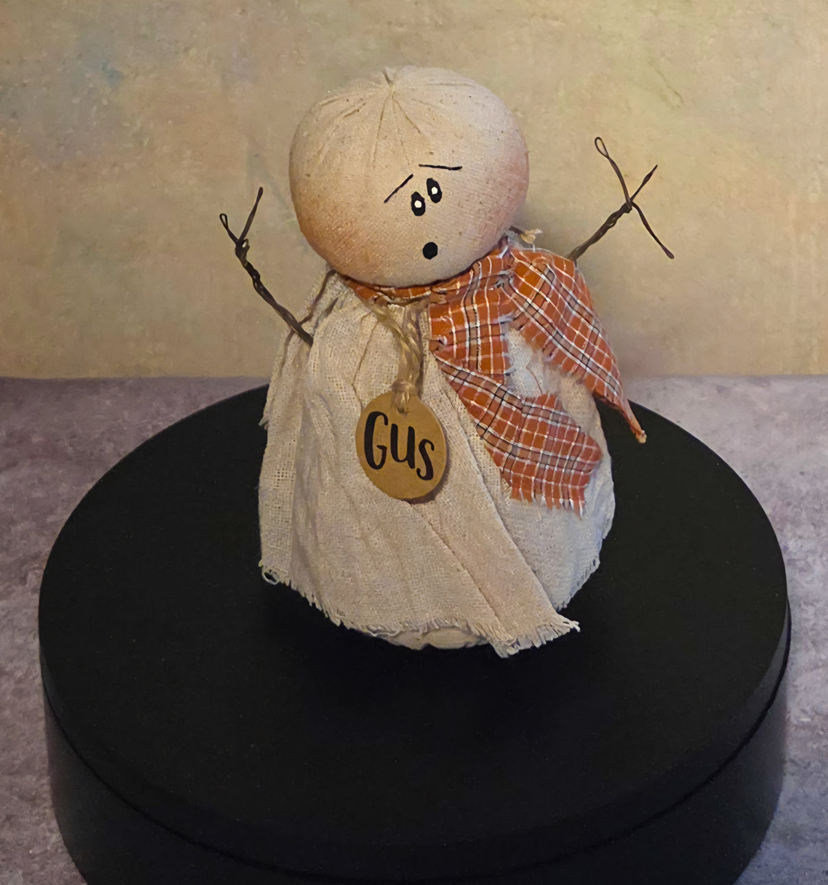 Front view of Gus ghost soft sculpture with distressed cream fabric, twig arms, orange plaid scarf, expressive face, and wooden name tag.