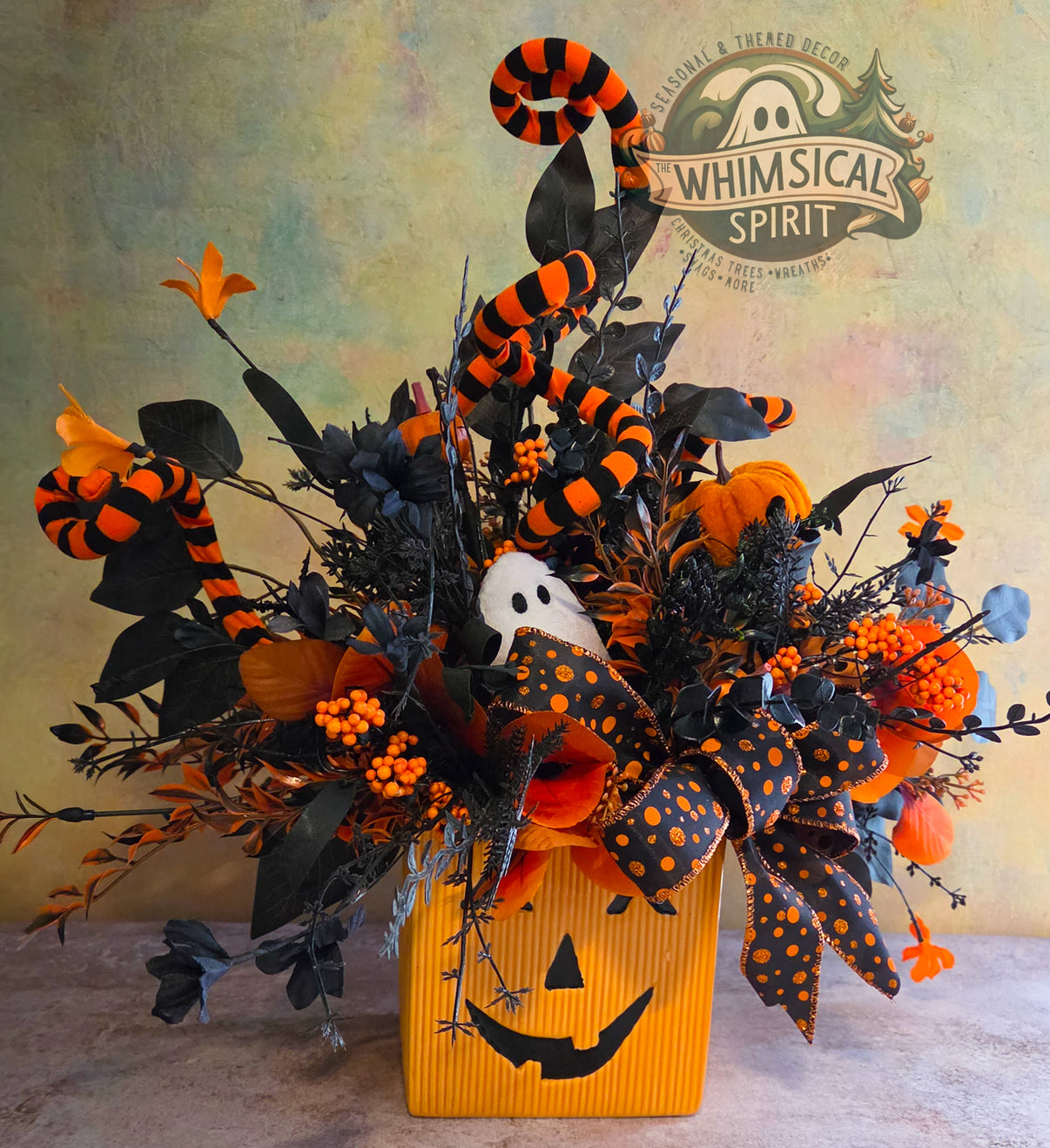 Front view of a jack-o’-lantern Halloween centerpiece with black foliage, orange flowers, striped spirals, and a ghost accent.