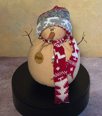 Front view of Jaxen snowman soft sculpture with a knit hat, red patterned scarf, carrot nose, twig arms, and wooden name tag.