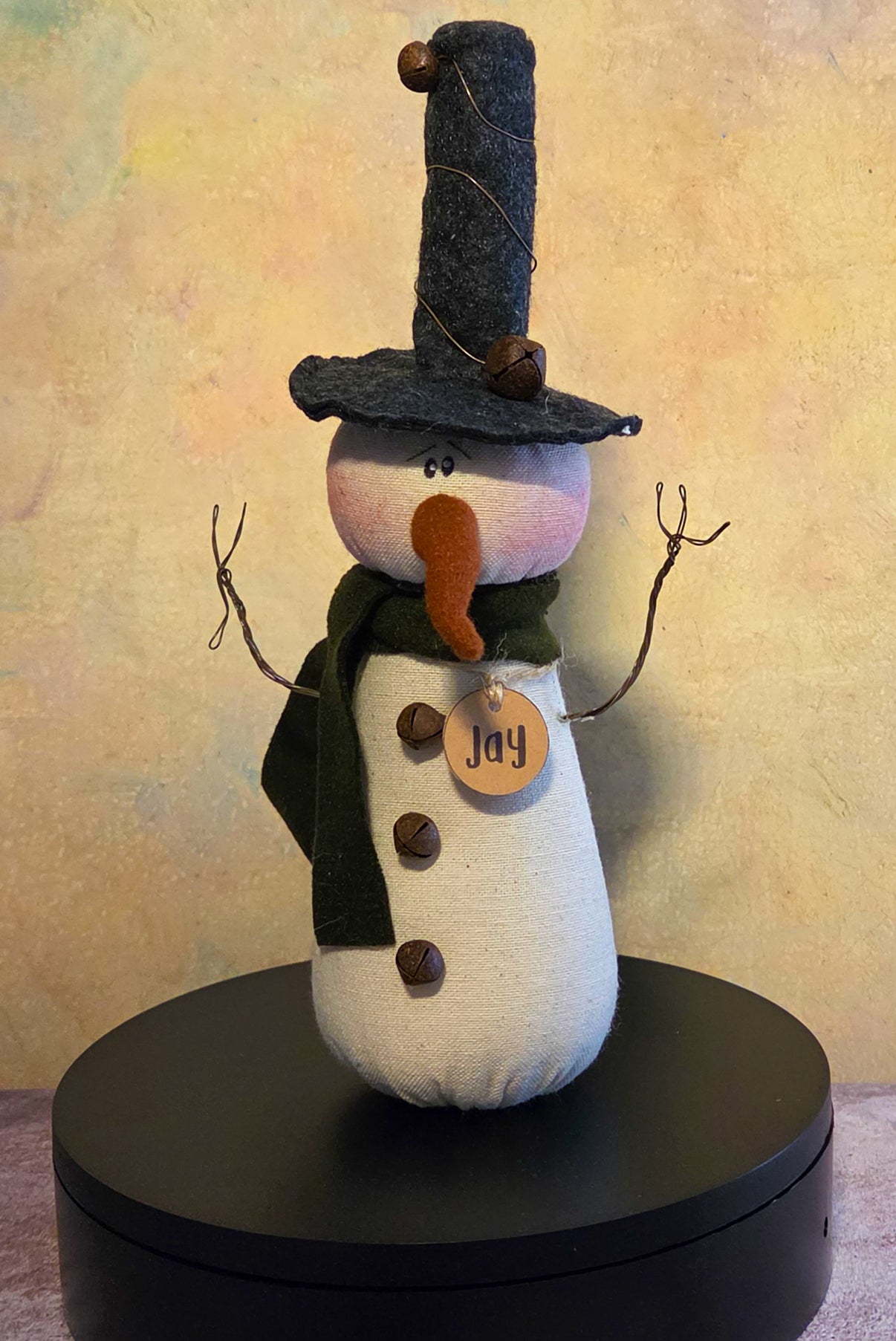 Jay the Snowman winter soft sculpture shelf sitter with tall black hat, green scarf, twig arms, carrot nose, button details, and name tag.