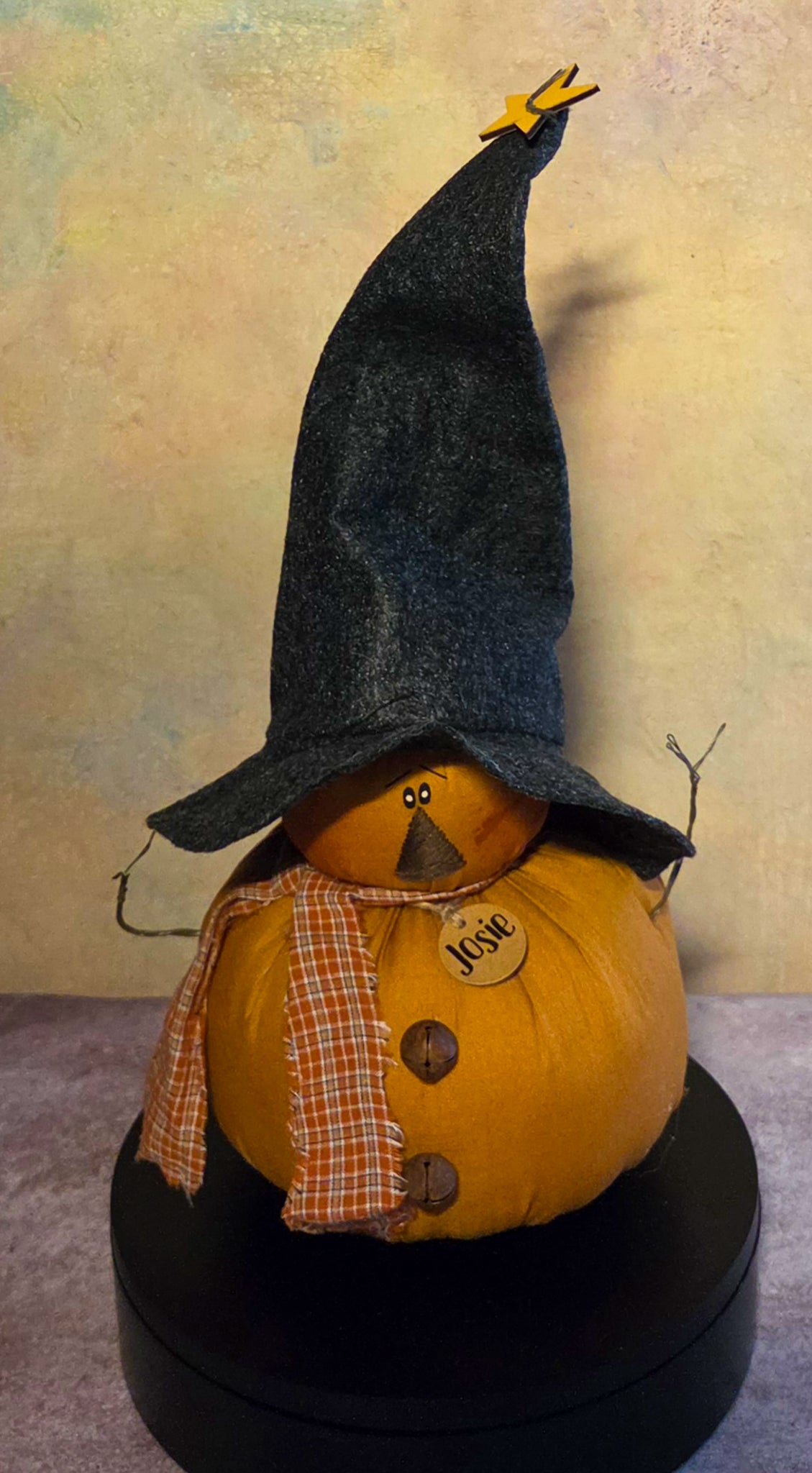 Front view of Josie pumpkin witch soft sculpture with a tall black hat, plaid scarf, twig arms, rustic buttons, and wooden name tag.