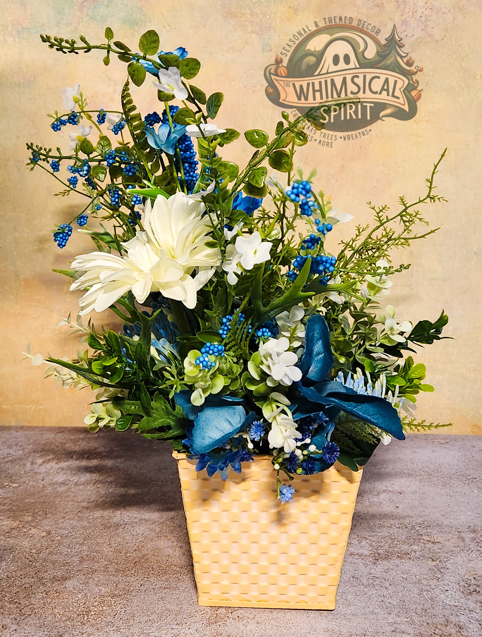 Floral arrangement in a woven basket with a blue ribbon on a textured surface, branded 'Whimsical Spirit'.
