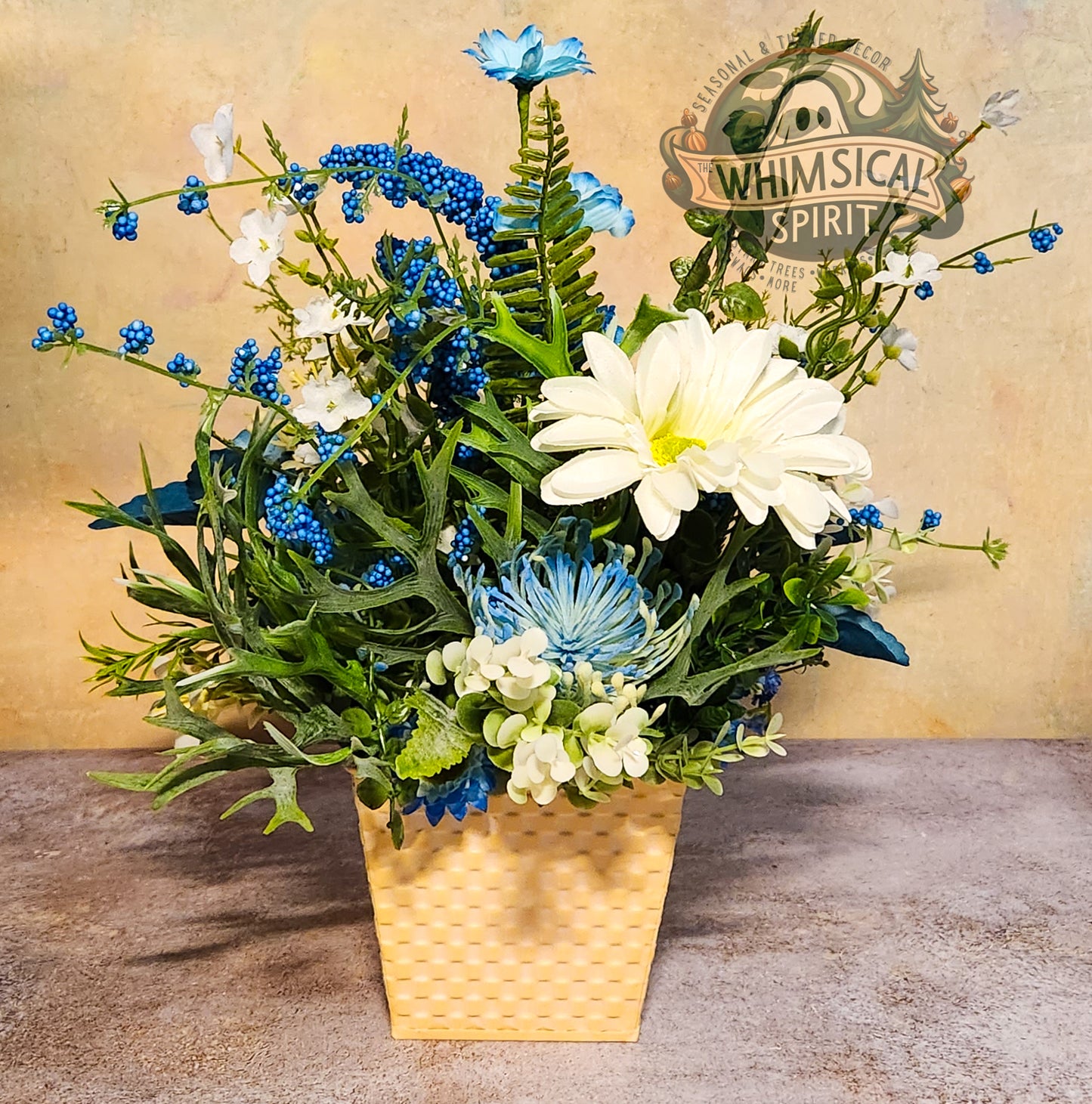 Artificial flower arrangement with blue and white flowers in a gold pot on a textured surface, branded 'Whimsical Spirit'.