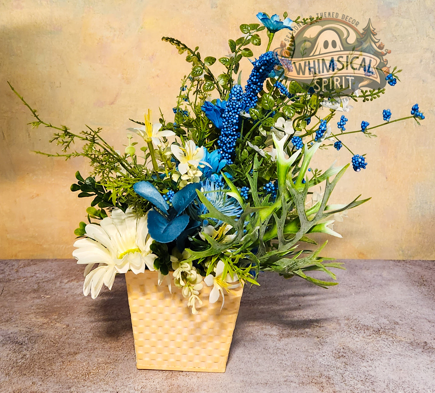 Floral arrangement with blue and green elements in a woven basket on a textured surface, featuring 'Whimsical Spirit' branding.
