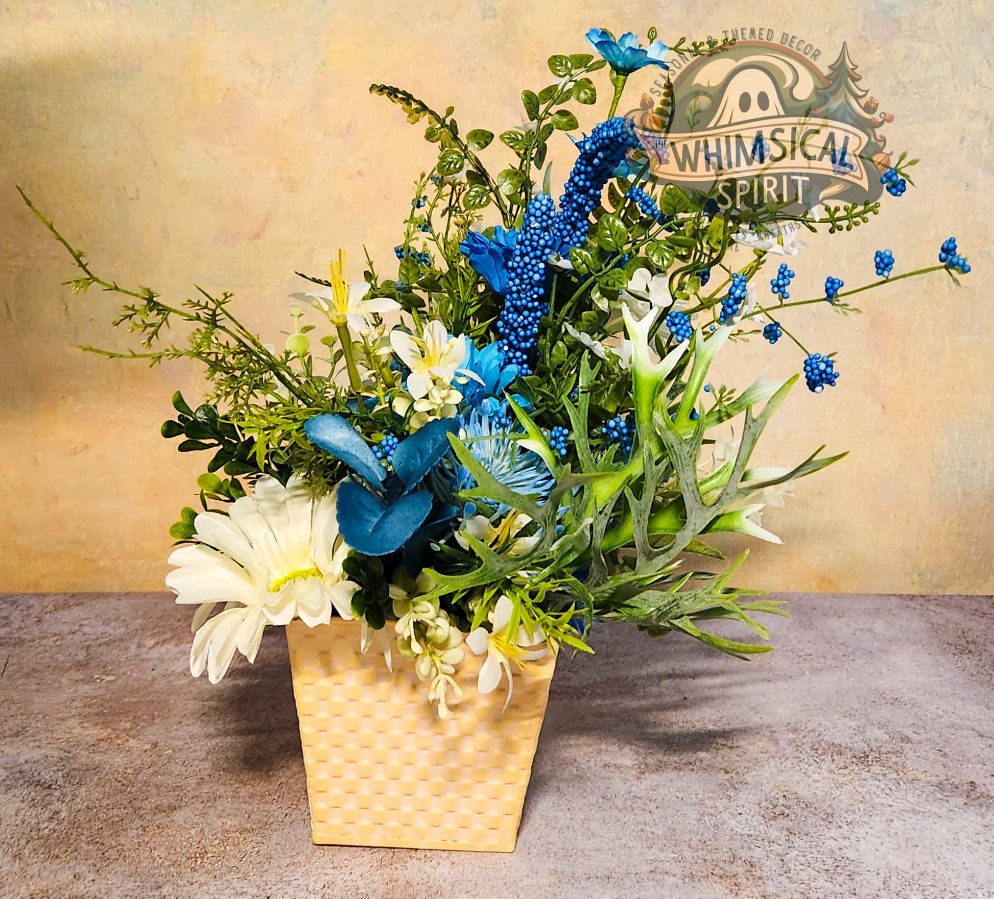 Floral arrangement with blue and green elements in a woven basket on a textured surface, featuring 'Whimsical Spirit' branding.