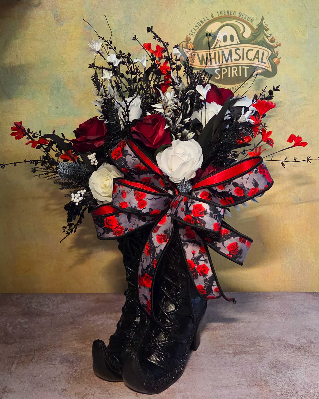 Handmade witch boot centerpiece with glossy black boots, red and white florals, black branches, and a large black-and-red rose ribbon bow.