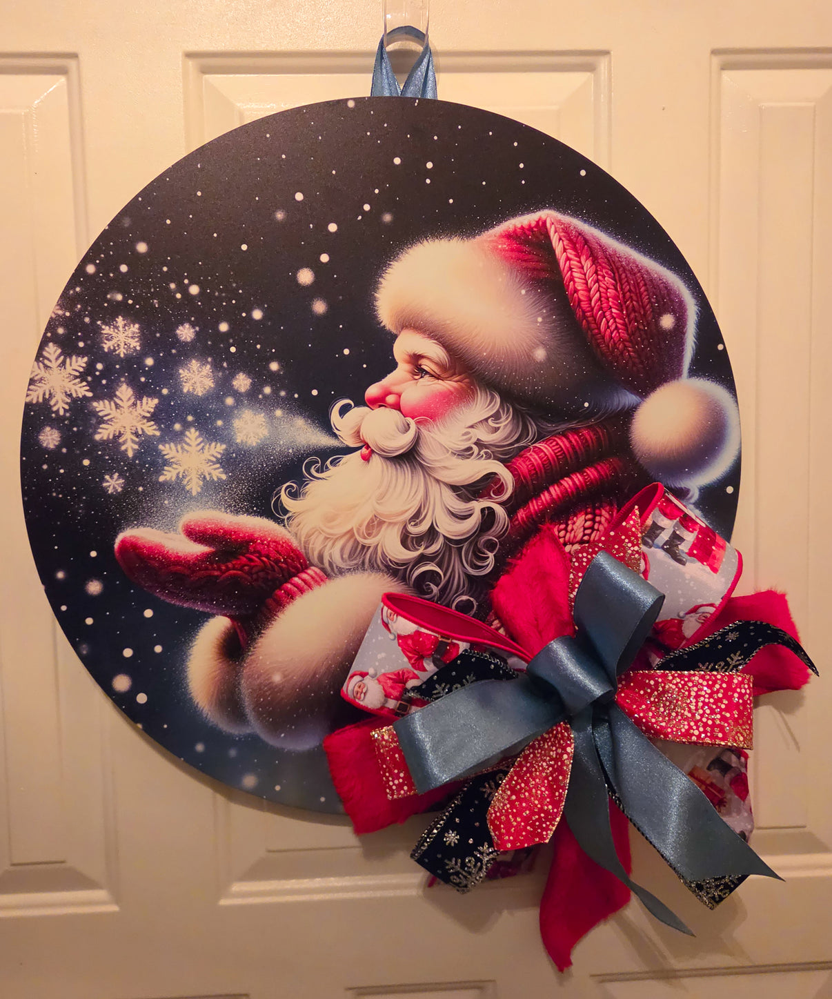 Midnight Snowfall Santa Christmas door hanger featuring Santa blowing snowflakes across a dark winter sky with a layered red, black, and gray bow.