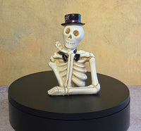 Small ivory skeleton resin figurine wearing a black top hat with purple trim and a black bow tie in a relaxed leaning pose.