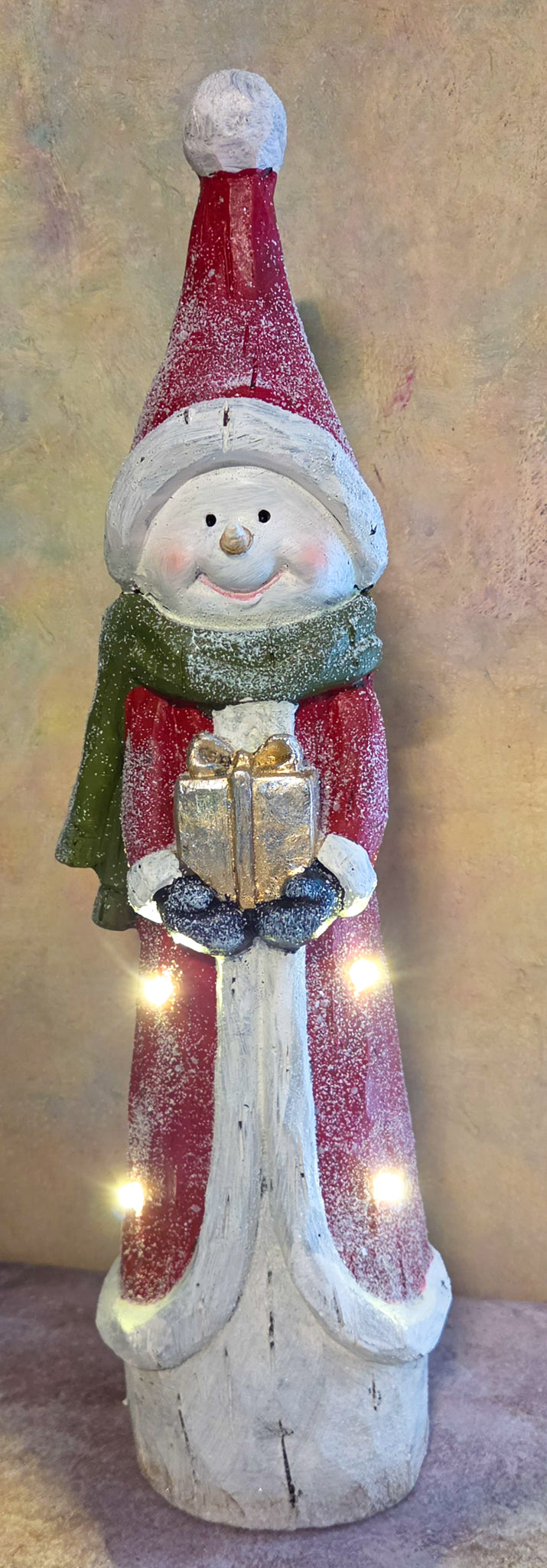 Front view of tall light-up snowman Christmas figurine wearing a red coat, green scarf, oversized Santa hat, and holding a gold present.