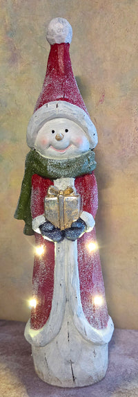 Front view of tall light-up snowman Christmas figurine wearing a red coat, green scarf, oversized Santa hat, and holding a gold present.